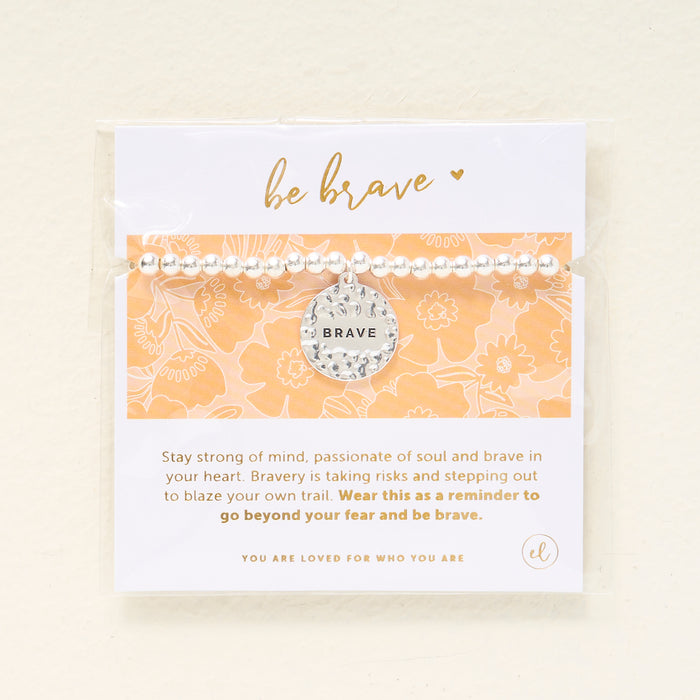 The Darling Effect Sentiment Beaded Charm Bracelet features silver beads and a round "BRAVE" charm, displayed on a yellow floral card that reads: be brave—wear this thoughtful gift as a reminder to go beyond your fear.