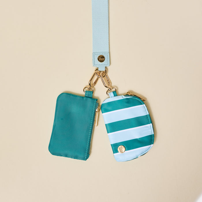 The Darling Effect Sicily Stripes Teal Dynamic Duo Pouch Wristlet features two small pouches—one solid teal, one in teal and white stripes—on a light blue strap with gold clasps, zippers, and a round gold emblem. Perfect for securing essentials.