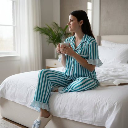 A woman wearing The Darling Effect's Sicily Stripes Teal Slumber Party Pajama Pants Set, crafted from ultra-soft fabric, sits on a made bed holding a mug and looking out the window. An open book and a green plant complete the cozy scene.