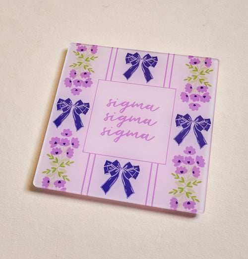 The Darling Effect's Sigma Sigma Sigma Acrylic Coaster features a pink and purple floral design with blue bows and "sigma" scripted three times—a perfect sorority gift for any occasion.