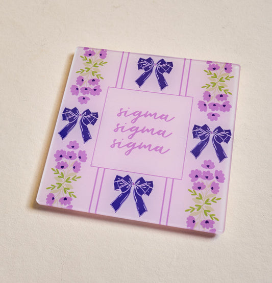 The Darling Effect's Sigma Sigma Sigma Acrylic Coaster features a pink and purple floral design with blue bows and "sigma" scripted three times—a perfect sorority gift for any occasion.