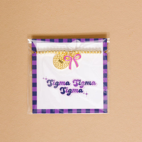 The Darling Effect's Sigma Sigma Sigma Bow Gold Charm Bracelet features a gold charm and pink bow, packaged with "Sigma Sigma Sigma" in purple script and a purple checkered border—a stylish way to show chapter pride.