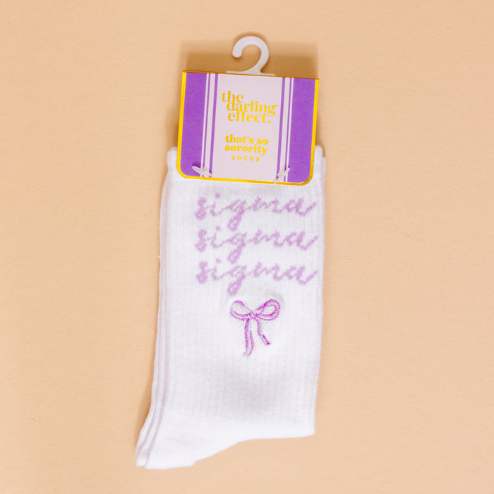 The Darling Effect’s Sigma Sigma Sigma Embroidered Crew Socks feature “sigma” in purple cursive three times with a small purple bow—displayed on peach background and packaging labeled “that’s so sorority socks”—perfect for sorority gifting.
