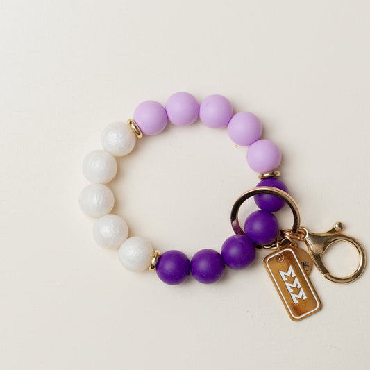 The Darling Effect’s Sigma Sigma Sigma Hands-Free Keychain Wristlet features white, lavender, and purple beads, a gold clasp, and a Greek letter charm tag.
