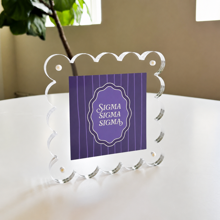 The Darling Effect’s Sigma Sigma Sigma Square Scalloped Acrylic Frame displays a purple “SIGMA SIGMA SIGMA” card on a white table, with a leafy plant and window in the background.