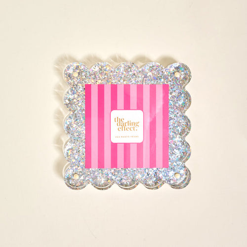 The Darling Effect’s Silver Glitter Square Scalloped Acrylic Picture Frame features a modern scalloped-edge design and holds an insert with pink and red stripes, displayed on a plain, light-colored background.