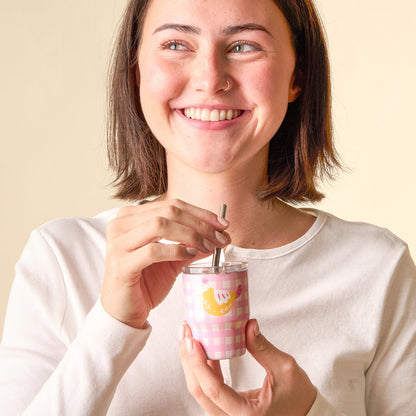 A young woman with short brown hair and a nose ring smiles while holding the-darling-effect Spring Chicken 2 oz Tiny Tumbler, a pink checkered cup featuring cute yellow bird prints and a metal straw. She wears a white long-sleeve shirt.