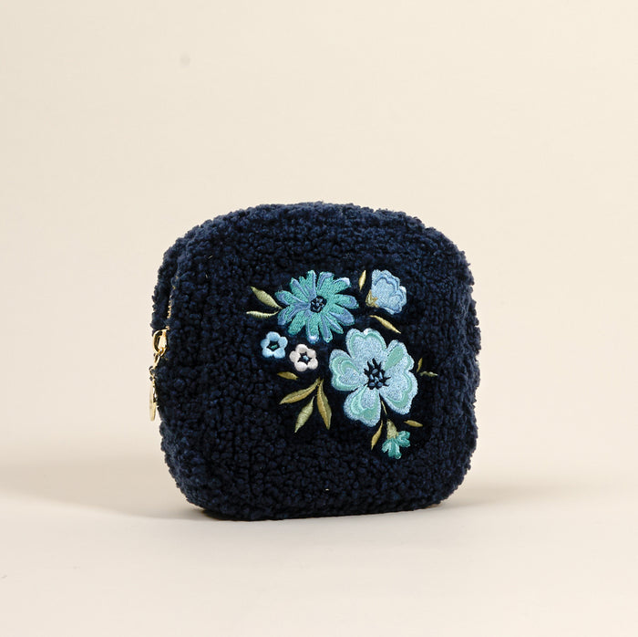 The Square Dainty Floral Navy Teddy Pouch by the-darling-effect is a navy blue sherpa cosmetic bag, adorned with embroidered blue and green flowers.
