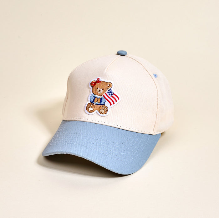 The Darling Effect's Star Spangled Bear Classic Canvas Hat features a beige crown, light blue brim, embroidered teddy bear with an American flag on the front, and a small blue button on top for a casual look.