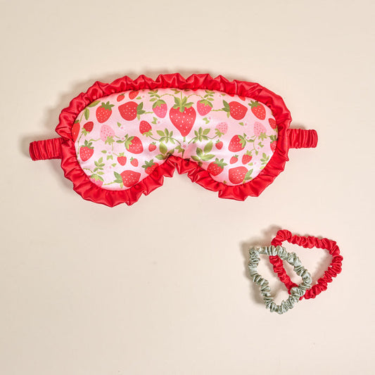 The Darling Effect’s Strawberry Fields Beauty Sleep Mask, featuring pink satin, red ruffles, and a strawberry print, pairs with matching red and light green satin scrunchies for a charming sleep set—shown against a beige background.