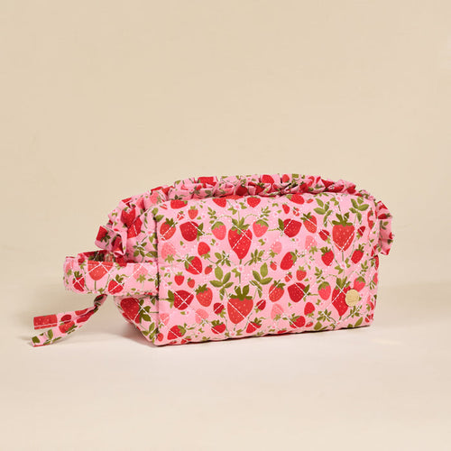 The Darling Effect’s Strawberry Fields Essentials Quilted Cosmetic Pouch features a pink quilted design with strawberry and leaf patterns, a ruffled top, and a side handle—ideal for travel—shown on a plain beige background.