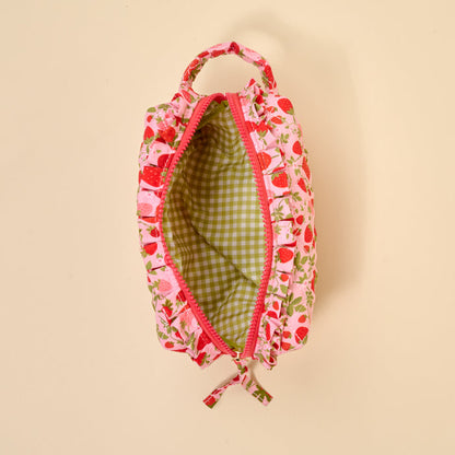 The Darling Effect Strawberry Fields Essentials Quilted Cosmetic Pouch features a pink strawberry print, red zipper, top handle, and green gingham lining, shown from above on a beige background.