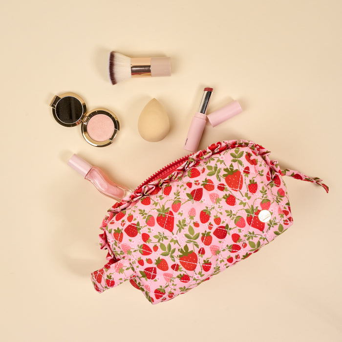 The Darling Effect's Strawberry Fields Essentials Quilted Cosmetic Pouch is open on a beige background, surrounded by a makeup brush, blending sponge, lipstick, blush compact, and nail polish.