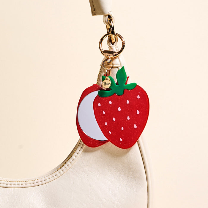 The Darling Effect’s Strawberry Mini Mirror Bag Charm, shaped like a strawberry with a gold ring, is attached to the handle of a white handbag, bringing playful charm to your accessories against a light beige background.