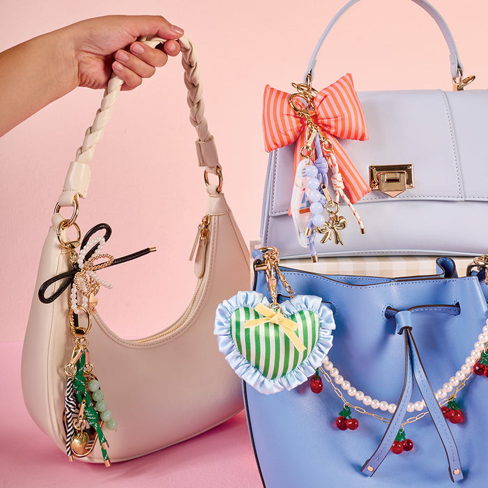 Three pastel purses are displayed with cute charms. The cream shoulder bag from The Darling Effect features a Strawberry Mini Mirror Bag Charm, while the light purple and blue bags are accented with playful, trendy bag accessories.