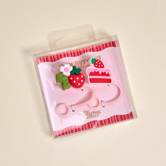 The Darling Effect’s Strawberry Shortcake Straw Topper Set features pink packaging with strawberry and cake designs. Includes three silicone toppers: two strawberries (with leaves/flower and leaf) and a cake slice—fun drinkware accessories!.