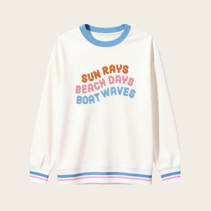 The Darling Effect’s Sun Rays, Beach Days, Boat Waves Oversized Crew features blue, pink, and orange text on beige with blue collar and cuffs detailed in pink and white stripes—a cozy wardrobe staple.
