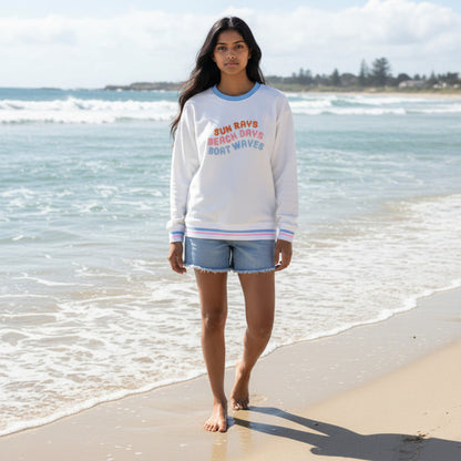 A woman with long dark hair walks barefoot on the sand, wearing The Darling Effect’s Sun Rays, Beach Days, Boat Waves Oversized Crew and denim shorts. Waves roll in behind her beneath a partly cloudy sky.