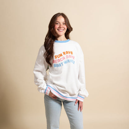 A smiling woman stands against a beige background wearing The Darling Effect’s Sun Rays, Beach Days, Boat Waves Oversized Crew—an oversized white sweatshirt with pastel trim and embroidered lettering, perfect for a cozy wardrobe.
