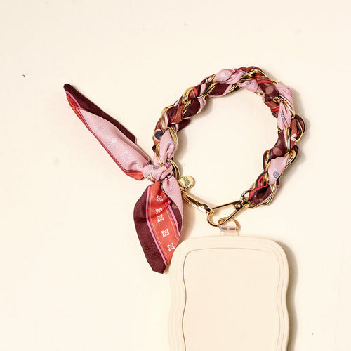The Darling Effect Sunset Tales Call Me Later Phone Scarf features a braided pink and burgundy silk scarf woven through a gold-tone chain, tied in a bow, and attached to a cream phone case with a gold clasp.