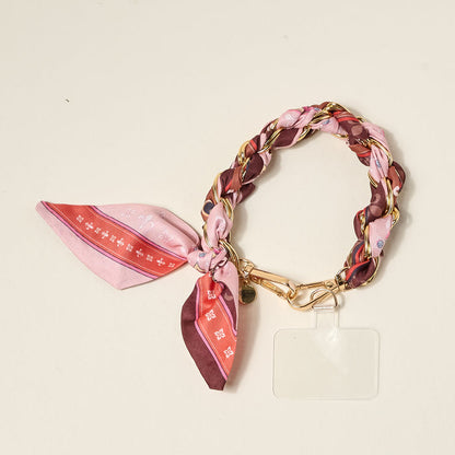 The Darling Effect’s Sunset Tales Call Me Later Phone Scarf features a pink and brown braided wristlet, gold-tone chain, and phone holder—a chic accessory for your device.