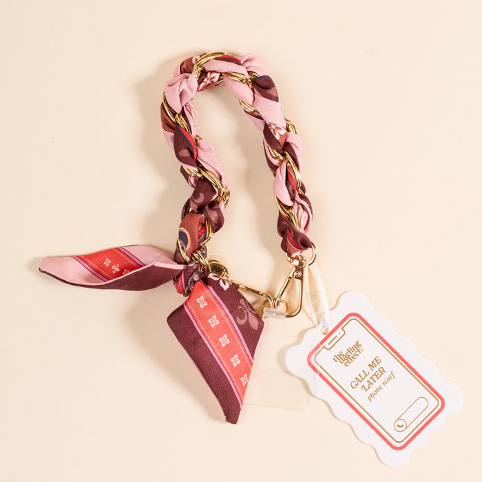 The Darling Effect's Sunset Tales Call Me Later Phone Scarf is a braided pink and gold-tone keychain with a clasp, phone scarf detail, and white CALL ME tag. Shown on a pale beige background.