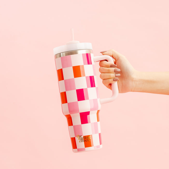 A hand holds The Darling Effect’s Sweetheart Check 40 oz Take Me Everywhere Tumbler, featuring a handle, straw, and custom pink, red, and white checkerboard design on a light pink background.