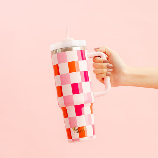 A hand holds The Darling Effect’s Sweetheart Check 40 oz Take Me Everywhere Tumbler, featuring a handle, straw, and custom pink, red, and white checkerboard design on a light pink background.