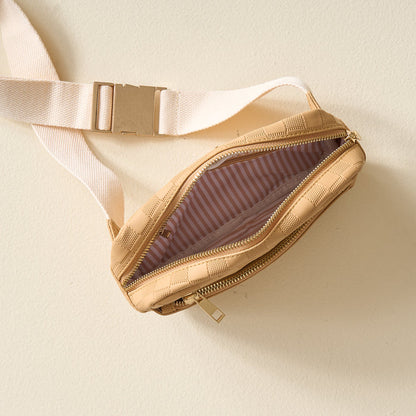 The Darling Effect’s Tan Urban Check Belt Bag, featuring beige checkered vegan leather, a cream strap, and gold buckle, sits unzipped on a light surface to reveal its pink and white striped fabric lining.