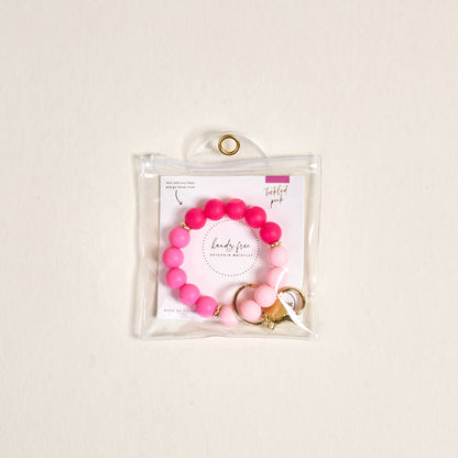 The Darling Effect Tickled Pink Hands-Free Keychain Wristlet, a beaded pink and white silicone key holder with gold accents, is shown in a clear pouch with a gold grommet. The pouch includes an insert reading "lady flow" and "Twisted pink.