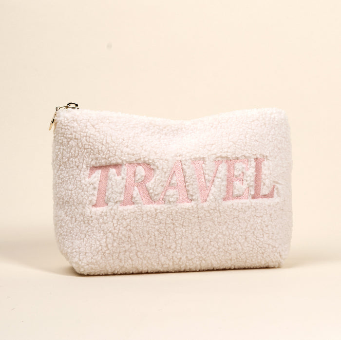 The Darling Effect’s Travel Edit Teddy Pouch is a fluffy white sherpa zippered pouch that stands upright, with “TRAVEL” boldly embroidered in large pink letters on the front.