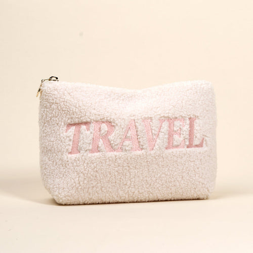 The Darling Effect’s Travel Edit Teddy Pouch is a fluffy white sherpa zippered pouch that stands upright, with “TRAVEL” boldly embroidered in large pink letters on the front.