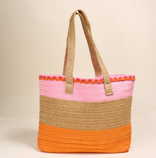 The Darling Effect's Tropical Sunset Straw & Sun Tote is a woven, lightweight bag with tan handles and horizontal tan, pink, and orange color blocks plus a pink trim—perfect for vacations and shown against a plain beige background.