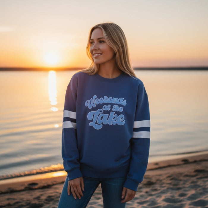 A woman in The Darling Effect’s Weekends at the Lake Oversized Crew smiles on a sandy lakeshore at sunset. The calm water and colorful sky highlight the cozy, relaxed vibe of this comfy sweatshirt.