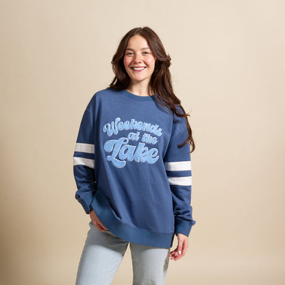 A smiling woman wears The Darling Effect’s Weekends at the Lake Oversized Crew, a blue sweatshirt with chenille lettering and white sleeve stripes, styled effortlessly with light wash jeans.
