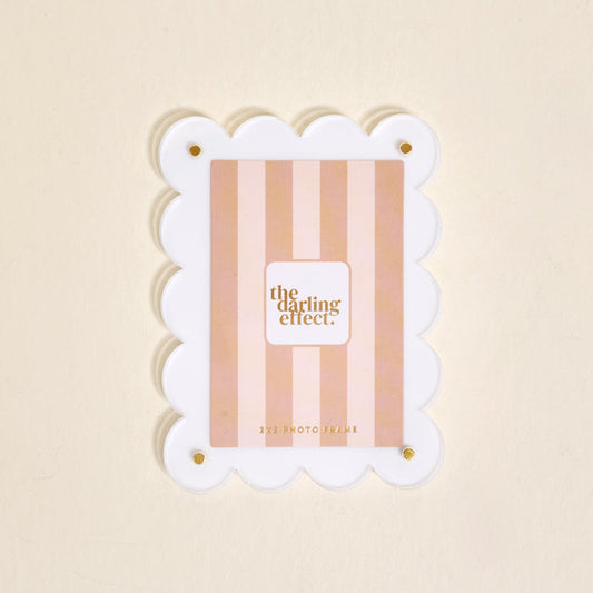 The Darling Effect White Mini Acrylic Picture Frame Magnet features scalloped white edges, gold corner screws, and a pink striped insert with the brand name. Shown against a light beige background.