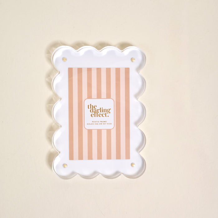 The White Rectangle Scalloped Acrylic Frame by the-darling-effect features a pink and beige striped insert with the brand name, displayed on a light cream background, adding a modern and charming touch to your decor.