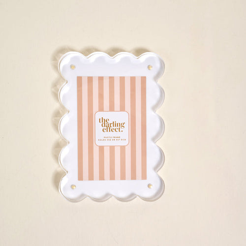 The White Rectangle Scalloped Acrylic Frame by the-darling-effect features a pink and beige striped insert with the brand name, displayed on a light cream background, adding a modern and charming touch to your decor.