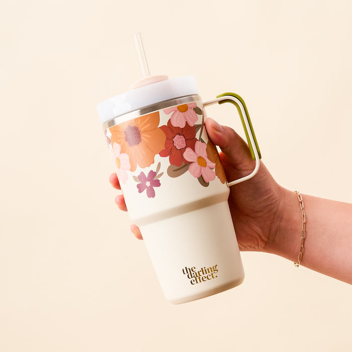 A hand holds a Wild About You Cream 20 oz Mini On-the-Go Tumbler by The Darling Effect, featuring pink and orange flowers, a green handle, white lid, and clear straw.