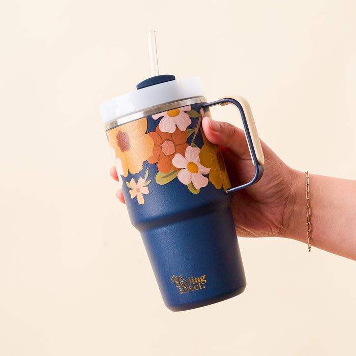 A hand holds The Darling Effect’s Wild About You Navy 20 oz Mini On-the-Go Tumbler, featuring a metal handle, white lid, clear straw, gold brand text near the base, and vibrant floral designs.