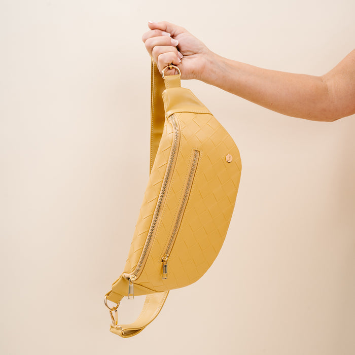 A hand displays The Darling Effect's Woven Mustard Trendy Luxe Belt Bag, a vegan leather accessory with gold hardware and zippered pocket—perfect for effortless, hands-free style against a beige backdrop.