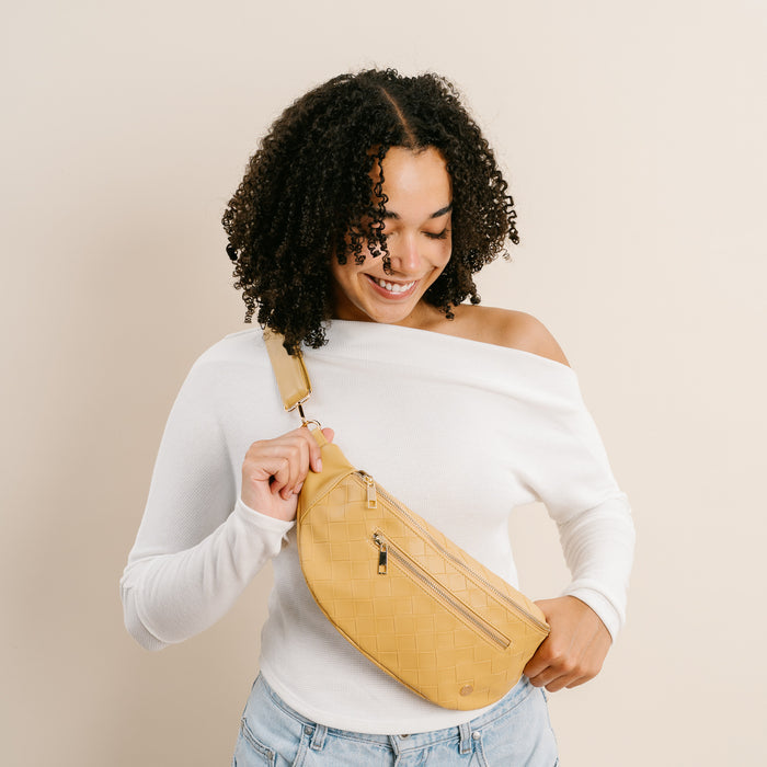 A smiling woman with curly hair models The Darling Effect's Woven Mustard Trendy Luxe Belt Bag in vegan leather, styled with a white off-shoulder top and light jeans for a chic, hands-free look against a beige background.