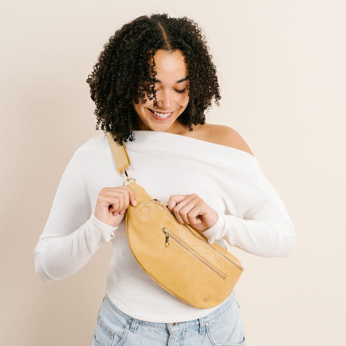 A person with curly hair, in a white off-shoulder top and light jeans, smiles while opening The Darling Effect’s Woven Mustard Trendy Luxe Belt Bag for a hands-free look against a plain beige background.