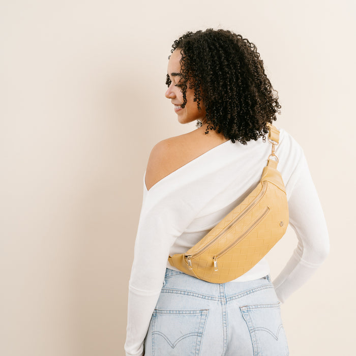 A woman with curly hair, in a white off-shoulder top and light blue jeans, stands facing away, looking over her shoulder. She carries The Darling Effect’s Woven Mustard Trendy Luxe Belt Bag across her back.