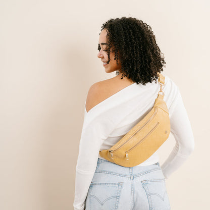 A woman with curly hair, in a white off-shoulder top and light blue jeans, stands facing away, looking over her shoulder. She carries The Darling Effect’s Woven Mustard Trendy Luxe Belt Bag across her back.