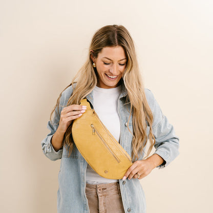 A woman with long blonde hair smiles, holding The Darling Effect's Woven Mustard Trendy Luxe Belt Bag in vegan leather. She pairs it with a light denim jacket, white shirt, and beige pants for a chic hands-free look against a light beige background.