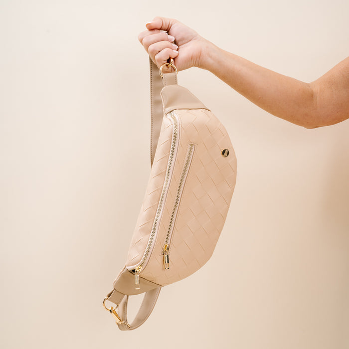 A hand holds The Darling Effect's Woven Oat Trendy Luxe Belt Bag, a beige vegan leather crossbody with gold zipper and hardware, set against a light beige background.