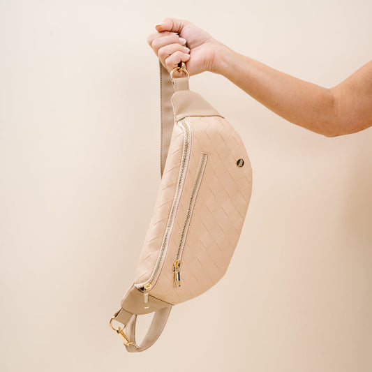 A hand holds The Darling Effect's Woven Oat Trendy Luxe Belt Bag, a beige vegan leather crossbody with gold zipper and hardware, set against a light beige background.