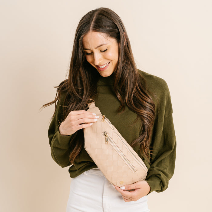 A woman with long brown hair, wearing an olive green sweater and white pants, smiles while holding and unzipping The Darling Effect's Woven Oat Trendy Luxe Belt Bag against a neutral background.