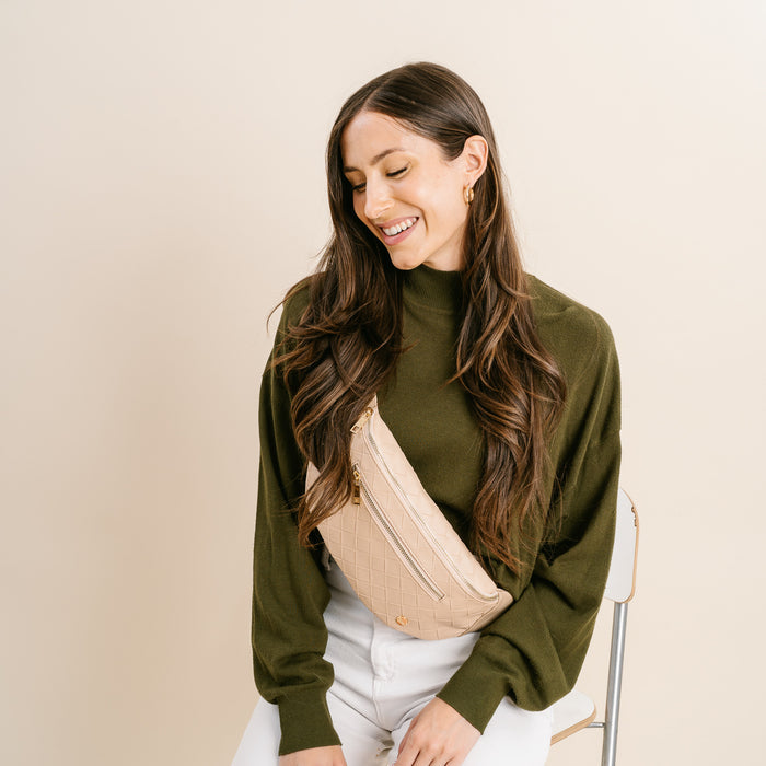 A woman with long brown hair in an olive green sweater and white pants smiles as she looks down, wearing The Darling Effect’s Woven Oat Trendy Luxe Belt Bag slung across her chest.